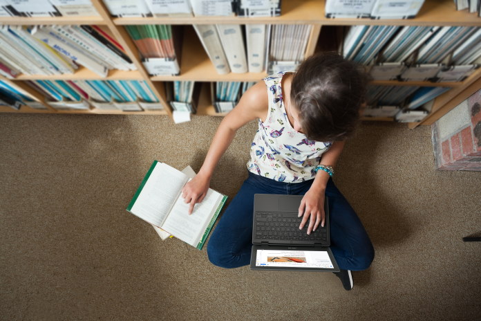 Girl working in a school library using a reference book and a laptop