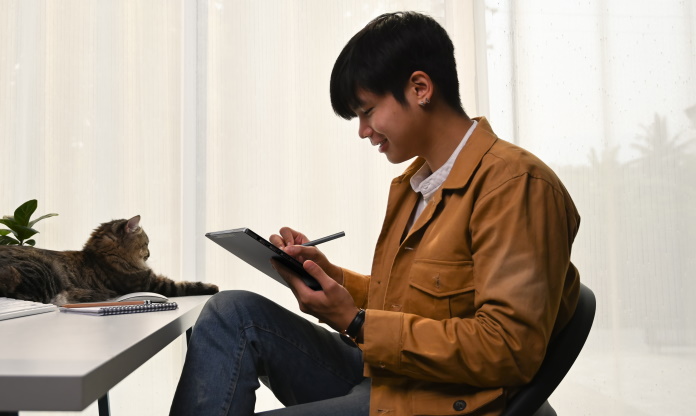 Student working on his laptop with a stylus while his cat sits nearby