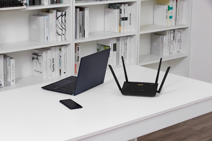 An ASUS laptop, router, and smartphone on a desk in an office with reference materials on shelves in the background