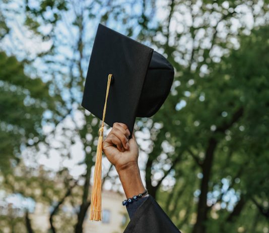 A young woman holding up a graduation hat into the air against a natural background of trees and sky