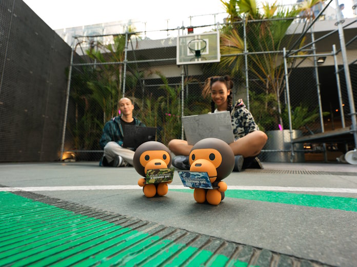 A young man and woman sitting on a basketball court with their Vivobook S 15 OLED BAPE Edition laptops with their Baby Milo accessory figurines in the foreground 