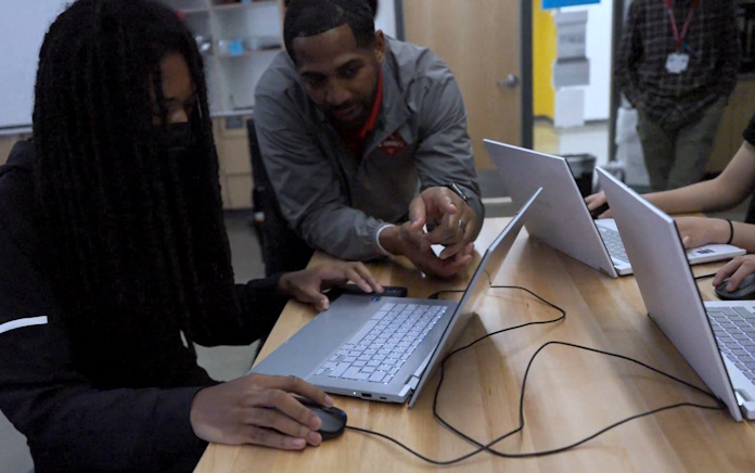 An instructor working with a student at a table filled with laptops