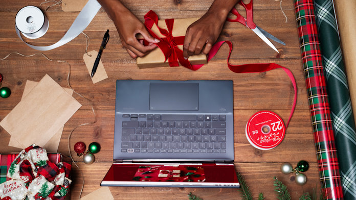 A person wrapping presents on a table full of paper, take, scissors, and other gift-wrapping elements in front of a Zenbook 14 OLED laptop