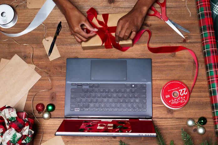A person wrapping up a present with brown paper and red ribbon on a wooden table in front of a Zenbook laptop