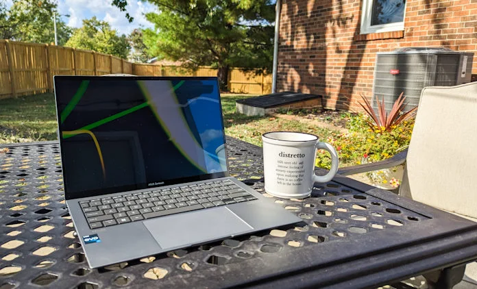 The Zenbook S 13 OLED laptop on a table with a coffee cup with the brick wall of the house in the background