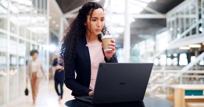A young woman hdoling a cup of coffee in a public space working on an ExpertBook B5 laptop