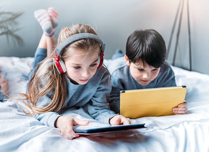 Two kids using devices while lying on a bed