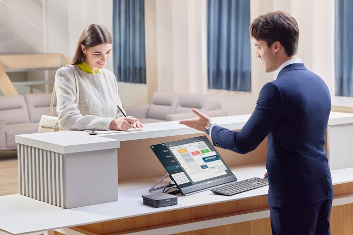A woman checks in at a reception desk equipped with a monitor and mini PC bundle